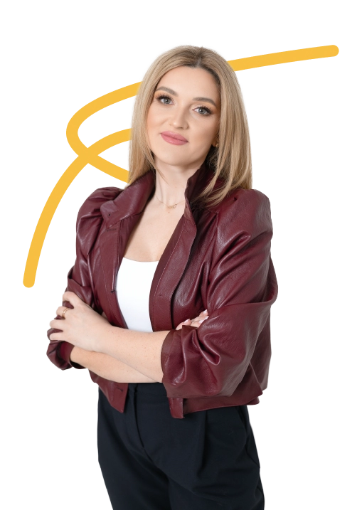 Blondynka w bordowej kurtce, białej bluzce i ciemnych spodniach, stoi ze skrzyżowanymi ramionami na niebieskim tle.